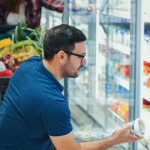 Person looking at container of dairy product, in front of a grocery store cooler